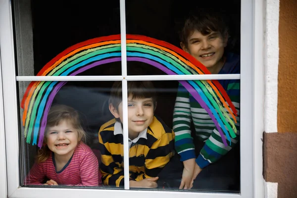 Tři děti, dva školáci a batole s duhou namalovanou barvou okna během pandemické koronavirové karantény. Děti malovat duhy se slovy Pojďme všichni být v pořádku. — Stock fotografie