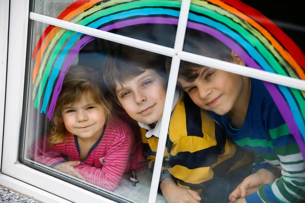 Drei Kinder, zwei Schulkinder Jungen und Kleinkind Mädchen mit Regenbogen bemalt mit bunten Fensterfarben während der Coronavirus-Quarantäne Pandemie. Kinder malen Regenbogen mit den Worten Lets all be well. — Stockfoto