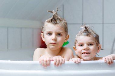 Mutlu kardeşler: İki küçük sağlıklı ikiz çocuk evde küvette banyo yaparak suyla birlikte oynuyorlar. Çocuklar birlikte eğleniyor. Çocuklar saçlarını şampuanla yıkıyor..