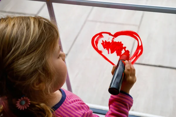 Gros plan de petite fille tout-petit avec coeur peint avec une couleur de fenêtre colorée pendant la quarantaine de coronavirus pandémique. Les enfants peignent des arcs-en-ciel dans le monde entier avec les mots "Soyons tous bien" . — Photo