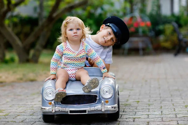 Zwei glückliche Kinder spielen mit einem großen alten Spielzeugauto im Sommergarten im Freien. Junge fährt Auto mit kleinem Kleinkind darin. Lachende und lächelnde Kinder. Familie, Kindheit, Lebensstil — Stockfoto