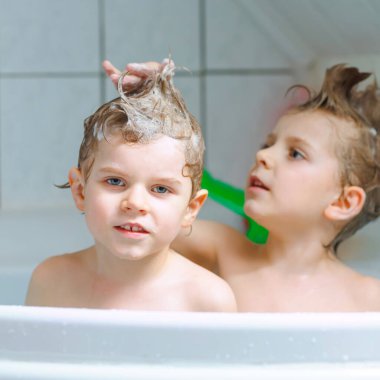 Mutlu kardeşler: İki küçük sağlıklı ikiz çocuk evde küvette banyo yaparak suyla birlikte oynuyorlar. Çocuklar birlikte eğleniyor. Çocuklar saçlarını şampuanla yıkıyor..