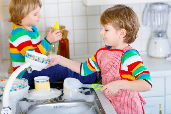 Dwóch małych blond chłopców zmywających naczynia w kuchni domowej. Dzieci, najlepsi przyjaciele dobrze się bawią pomagając w pracy domowej. Wewnątrz, rodzeństwo w kolorowych ubraniach — Zdjęcie stockowe