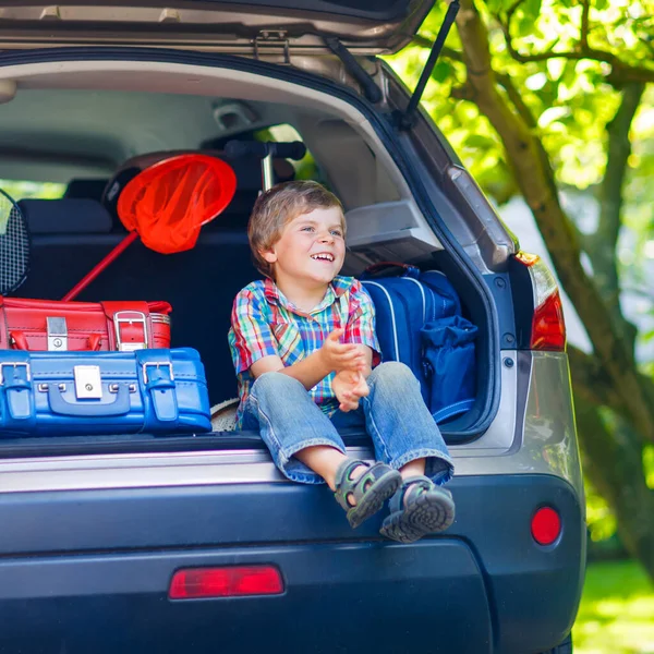 Menino animado sentado no porta-malas do carro pouco antes de sair para férias de verão com sua família. Criança feliz com malas e brinquedos em viagem . — Fotografia de Stock