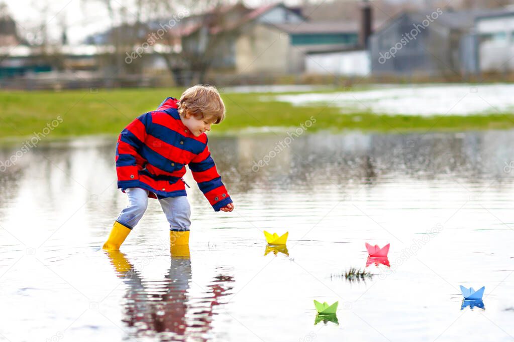 Happy little kid boy in yellow rain boots playing with paper ship boat by huge puddle on spring or autumn day. Active leisure for children. Funny child having fun outdoors, wearing colorful clothes.