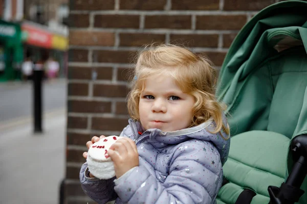 Menina pequena bonito sentado em carrinho de bebê enquanto caminhava com os pais nas ruas da cidade grande. Criança ativa feliz comendo bolo grande . — Fotografia de Stock