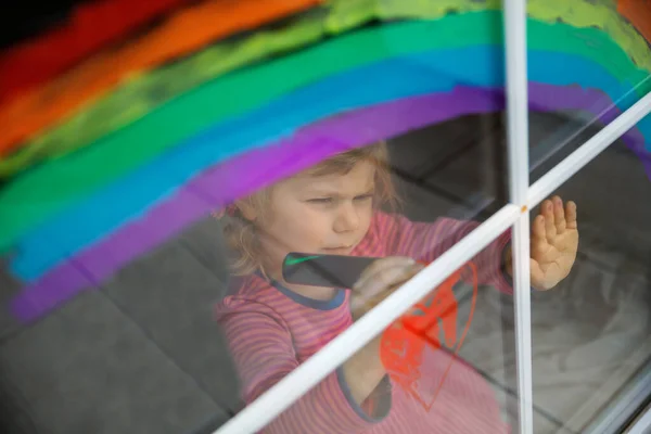 Adoralbe kleine peuter meisje met regenboog geschilderd met kleurrijke venster kleur tijdens pandemische coronavirus quarantaine. Kind schilderen regenbogen over de hele wereld met de woorden Laten we allemaal goed zijn. — Stockfoto