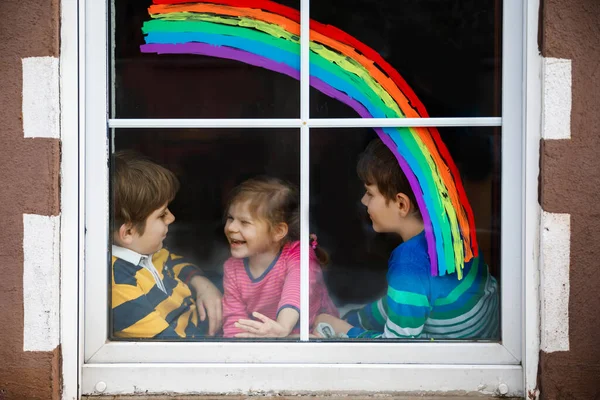 Drei Kinder, zwei Schulkinder Jungen und Kleinkind Mädchen mit Regenbogen bemalt mit bunten Fensterfarben während der Coronavirus-Quarantäne Pandemie. Kinder malen Regenbogen mit den Worten Lets all be well. — Stockfoto