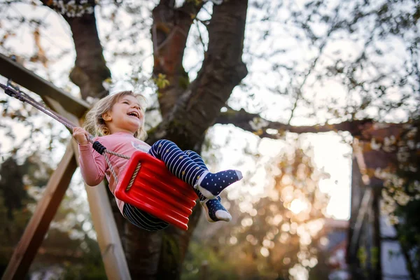 Glad vacker liten småbarn flicka som har kul på swing i hemmets trädgård. Söta friska barn svänger under blommande träd på soliga vårdagen. Baby skrattar och gråter — Stockfoto