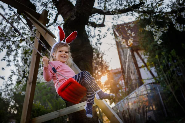 Šťastné krásné malé batole holčička s červenými velikonočními zajíčky uši baví na houpačce v domácí zahradě. Roztomilé zdravé dítě houpající se pod kvetoucími stromy za slunečného jarního dne. Dětský smích a pláč — Stock fotografie
