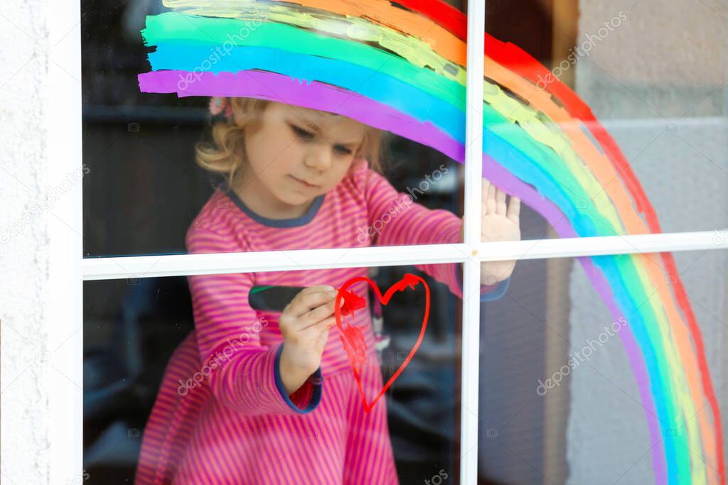 Adoralbe little toddler girl with rainbow painted with colorful window color during pandemic coronavirus quarantine. Child painting rainbows around the world with the words Lets all be well.