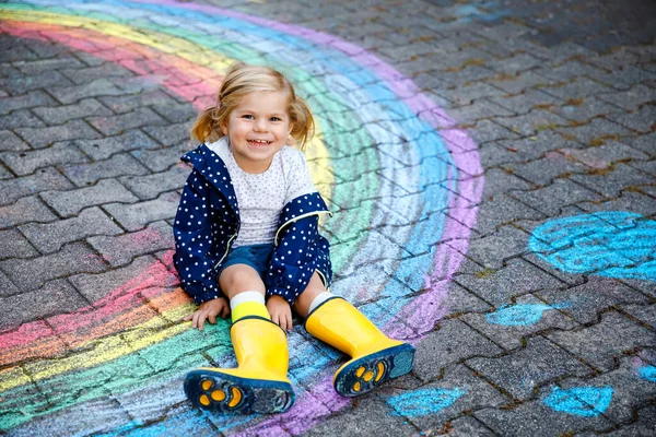 Gelukkige kleine peuter meisje in rubberen laarzen met regenboog geschilderd met kleurrijke krijt op de grond tijdens pandemische coronavirus quarantaine. Kinderen schilderen regenbogen samen met de woorden Laten we allemaal goed zijn — Stockfoto