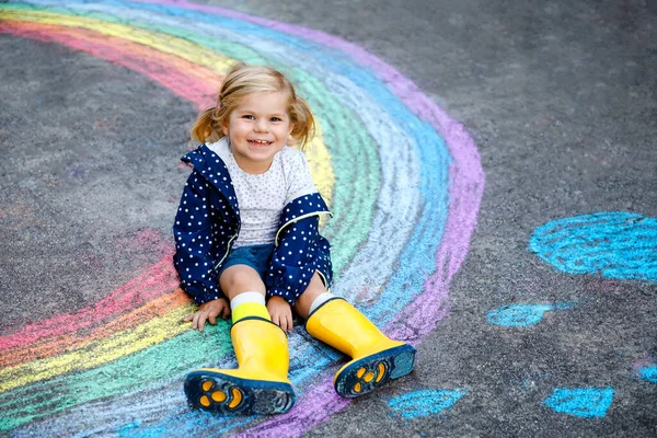 Gelukkige kleine peuter meisje in rubberen laarzen met regenboog geschilderd met kleurrijke krijt op de grond tijdens pandemische coronavirus quarantaine. Kinderen schilderen regenbogen samen met de woorden Laten we allemaal goed zijn — Stockfoto