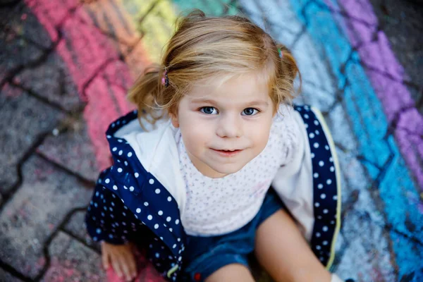 Pequeña niña feliz en botas de goma con sol de arco iris y nubes con lluvia pintadas con tiza de colores en el suelo o asfalto en verano. Lindo niño divirtiéndose. ocio creativo — Foto de Stock