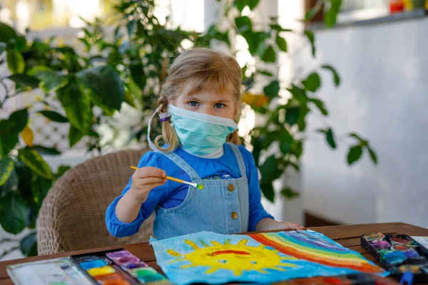 Маленькая девочка в медицинской маске рисует радугу акварелью во время пандемии коронавируса. Дети рисуют радуги по всему миру словами "Давайте все будем здоровы" . — стоковое фото