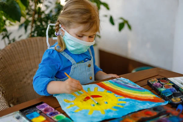 little toddler girl in medical mask painting rainbow with water colors during pandemic coronavirus quarantine disease. Children painting rainbows around the world with the words Lets all be well.