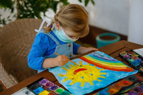 Klein peuter meisje in medisch masker schilderen regenboog met waterverf tijdens pandemische coronavirus quarantaine ziekte. Kinderen schilderen regenbogen over de hele wereld met de woorden Laten we allemaal goed zijn. — Stockfoto