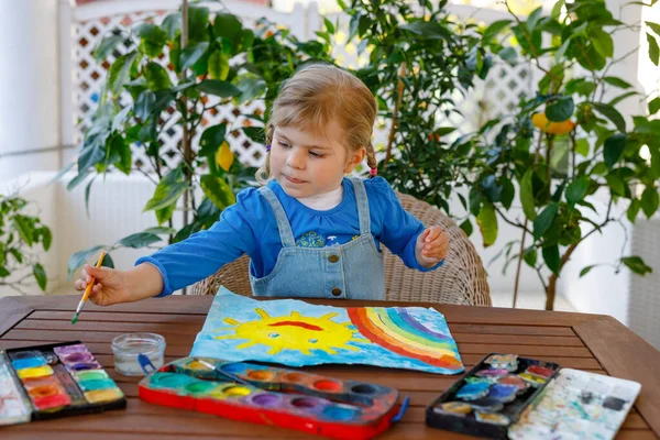 在大流行的验尸检疫期间，幼儿小女孩用水彩画彩虹和太阳。世界各地的孩子都在画彩虹，上面写着："让我们好好生活吧！"快乐的孩子 — 图库照片