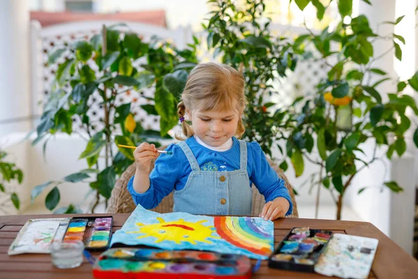 Little toddler girl painting rainbow and sun with water colors during pandemic coronavirus quarantine disease. Children painting rainbows around the world with the words Lets all be well. Happy child — Stock Photo, Image