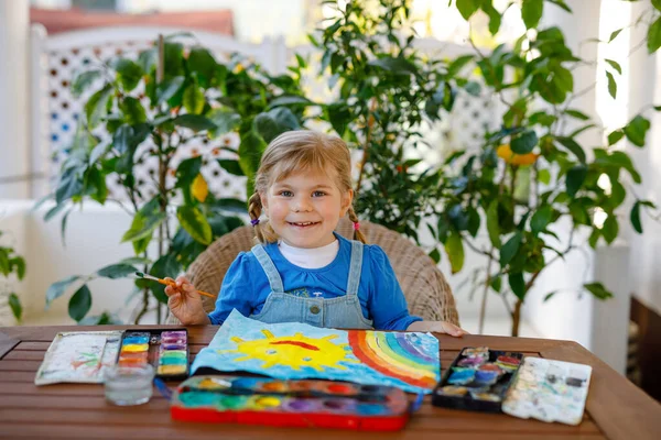 Petite fille tout-petit peignant arc-en-ciel et soleil avec des aquarelles pendant la maladie de quarantaine de coronavirus pandémique. Les enfants peignent des arcs-en-ciel dans le monde entier avec les mots "Soyons tous bien. Joyeux enfant — Photo