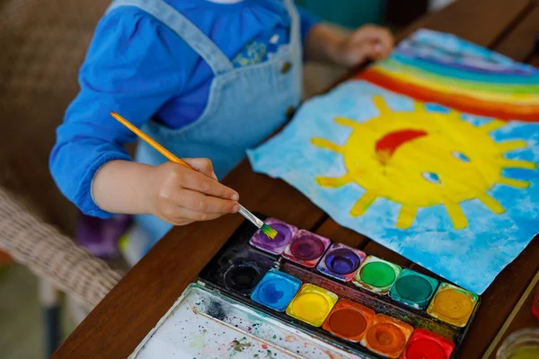 Pequeña niña pintando arco iris y sol con acuarelas durante la enfermedad pandémica de cuarentena por coronavirus. Niños pintando arcoíris alrededor del mundo con las palabras Vamos a estar todos bien. Niño feliz. — Foto de Stock