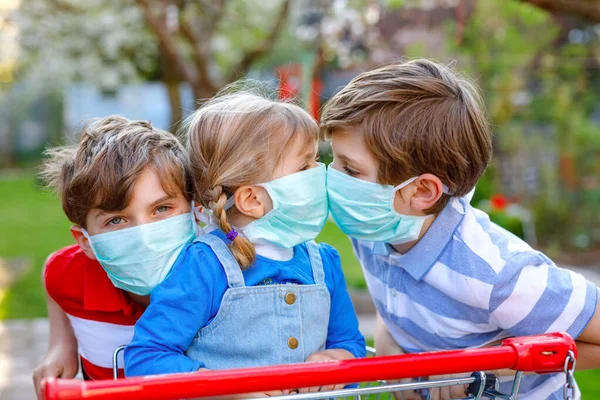 Três crianças, uma criança e dois miúdos mascarados como protecção contra a doença pandémica do coronavírus. Crianças com carrinho de compras usando equipamentos de proteção como luta contra o covid 19. — Fotografia de Stock