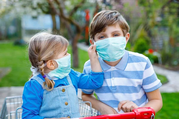 Dos niños, la niña y el niño de la escuela en máscara médica como protección contra la enfermedad pandémica por coronavirus. Niños hermanos con carrito de la compra utilizando equipo de protección como lucha con covid 19. — Foto de Stock