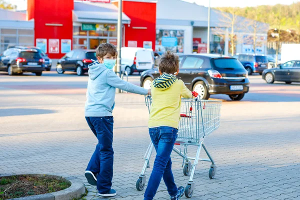 Two kids boys in medical mask as protection against pandemic coronavirus disease. Children using protective equipment against covid 19 and going for shopping in supermarket with cart trolley.