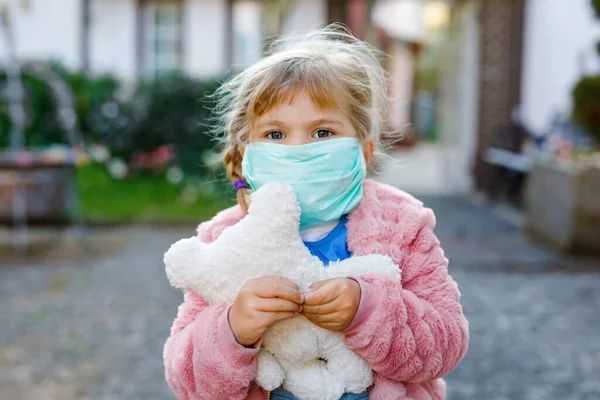 Menina pequena em máscara médica como proteção contra a doença de quarentena do coronavírus pandêmico. Criança bonito usando equipamentos de proteção como luta contra covid 19 e segurando urso brinquedo. — Fotografia de Stock