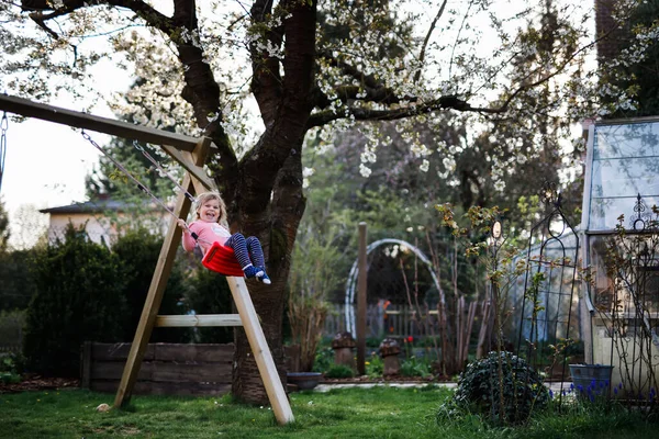 Menina pequena bonita feliz da criança que se diverte no balanço no jardim doméstico. Criança saudável bonito balançando sob árvores florescendo no dia ensolarado da primavera. Bebê rindo e chorando — Fotografia de Stock