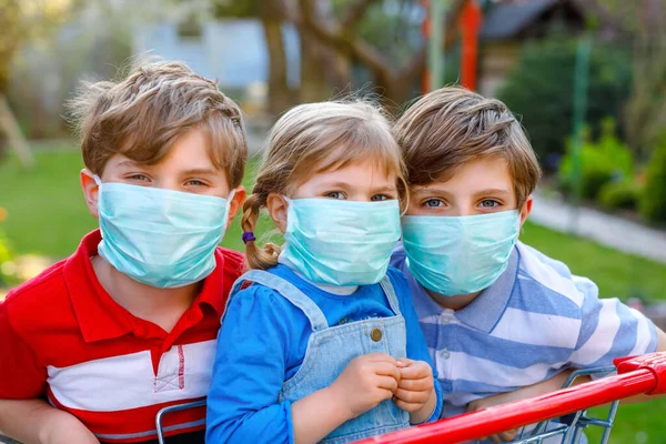 Três crianças, uma criança e dois miúdos mascarados como protecção contra a doença pandémica do coronavírus. Crianças com carrinho de compras usando equipamentos de proteção como luta contra o covid 19. — Fotografia de Stock