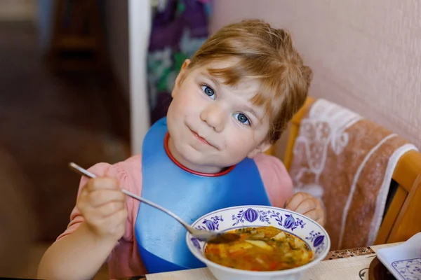 Adorável menina criança comendo sopa de legumes cozidos frescos na cozinha. Criança feliz come comida saudável ao almoço ou jantar. Aprendizagem de bebés. Casa, berçário, escola infantil ou creche — Fotografia de Stock