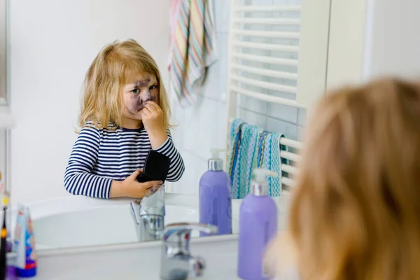 Funny little toddler girl using mothers make up and painting face with eye shadows. Happy baby child making experiments with cosmetics of mother. Kid around — Stock Photo, Image