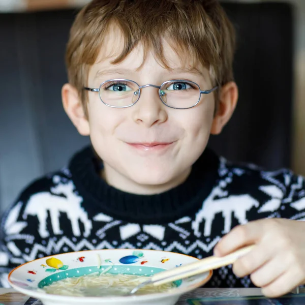 Menino da escola comendo sopa de legumes no interior. Criança loira na cozinha doméstica ou na cantina da escola. Criança bonito e comida saudável, sopa vegan orgânica com macarrão, frango e legumes . — Fotografia de Stock