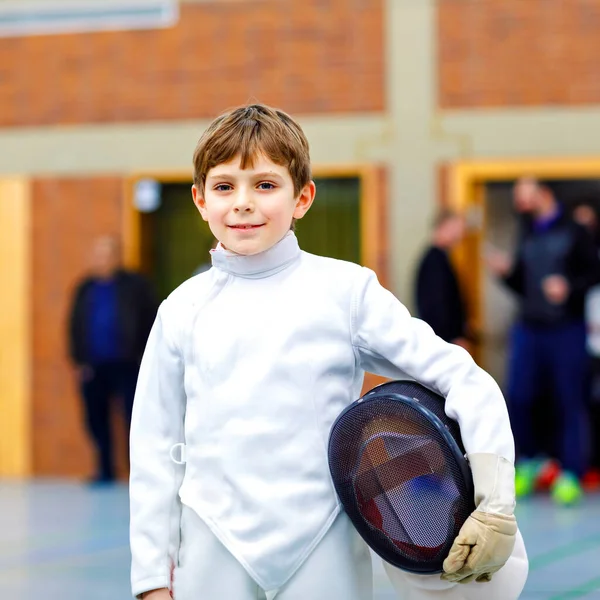 Malý chlapec šermuje na šermířské soutěži. Dítě v bílé šermířské uniformě s maskou a šavlí. Aktivní trénink dětí s učitelem a dětmi. Zdravé sporty a volný čas. — Stock fotografie