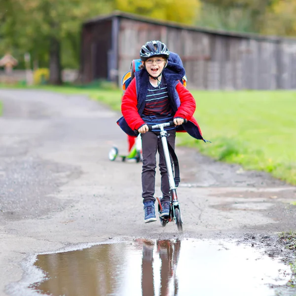 Menino da escola pequeno bonito montando em empurrar scooter no caminho para ou da escola. Estudante de 7 anos dirigindo através da poça de chuva. criança feliz engraçada em roupas de moda coloridas e com capacete. — Fotografia de Stock