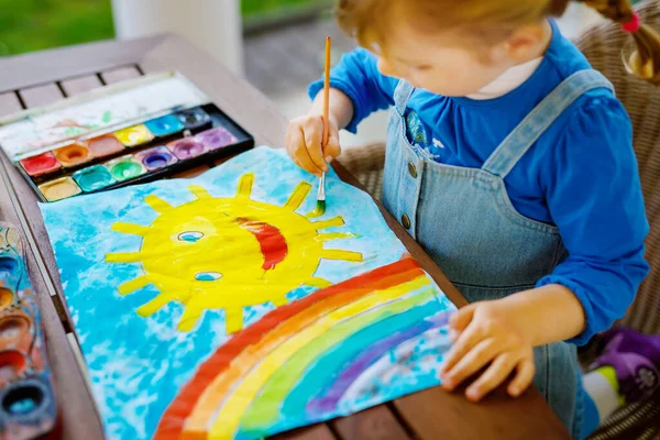 Liten småbarn flicka måla regnbåge och sol med akvareller under pandemisk coronavirus karantän sjukdom. Barn måla regnbågar runt om i världen med orden Låt oss alla vara bra. Lyckligt barn — Stockfoto