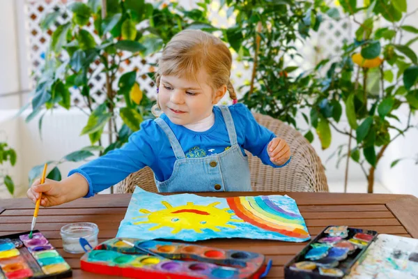 Petite fille tout-petit peignant arc-en-ciel et soleil avec des aquarelles pendant la maladie de quarantaine de coronavirus pandémique. Les enfants peignent des arcs-en-ciel dans le monde entier avec les mots "Soyons tous bien. Joyeux enfant — Photo