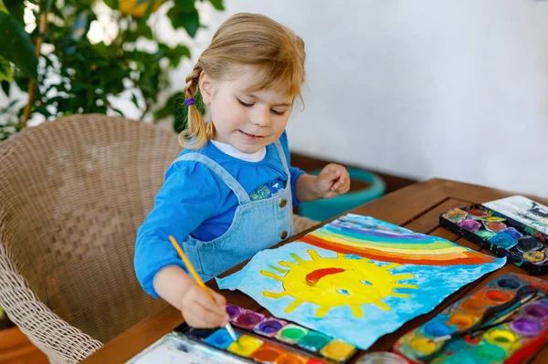 在大流行的验尸检疫期间，幼儿小女孩用水彩画彩虹和太阳。世界各地的孩子都在画彩虹，上面写着："让我们好好生活吧！"快乐的孩子 — 图库照片