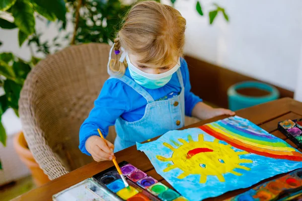 Pequena menina na máscara médica pintando arco-íris com cores de água durante a pandemia coronavírus quarentena doença. Crianças pintando arco-íris ao redor do mundo com as palavras Lets all be well . — Fotografia de Stock