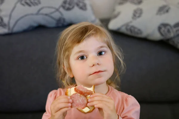 Adorable little toddler girl eating fresh prepared salami sandwith in kitchen. Happy child eats healthy food for lunch or dinner. Baby learning. Home, nursery, playschool or daycare — Stock Photo, Image