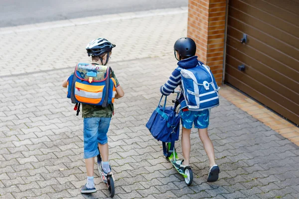 在阳光明媚的日子里，两个戴着安全帽的小学生带着背包在城市里骑着摩托车。穿着五颜六色衣服的快乐孩子在上学的路上骑自行车。从背面上方无法识别的面 — 图库照片