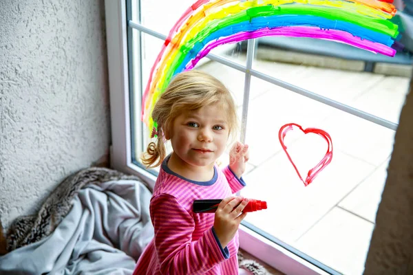Adoralbe bambina con arcobaleno dipinto con colorato colore della finestra durante la quarantena pandemica coronavirus. Bambino pittura arcobaleni in tutto il mondo con le parole Lascia che tutto sia bene. — Foto Stock