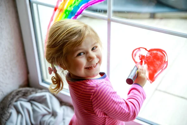 Adoralbe kleine peuter meisje met regenboog geschilderd met kleurrijke venster kleur tijdens pandemische coronavirus quarantaine. Kind schilderen regenbogen over de hele wereld met de woorden Laten we allemaal goed zijn. — Stockfoto