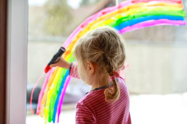 Adoralbe little toddler girl with rainbow painted with colorful window color during pandemic coronavirus quarantine. Child painting rainbows around the world with the words Lets all be well. — Stock Photo, Image