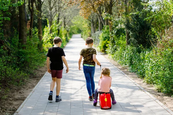 Trójka dzieci, mała dziewczynka i dwóch chłopców spacerujących w parku w chorobie koronawirusowej. Dzieci, piękne rodzeństwo bawiące się razem jako rodzina, używając walizki z zabawkami na kółkach — Zdjęcie stockowe