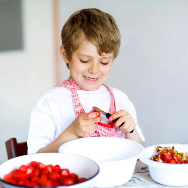 Little blond kid boy helping and making strawberry jam in summer. Funny child cleaning berries and preparing for cooking jam. Kid eating ripe strawberries in domestic kitchen — Stock Photo, Image