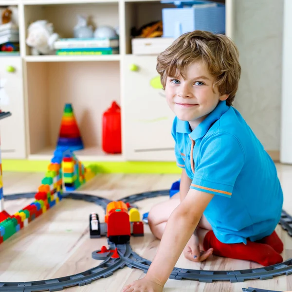 Adorable niño rubio jugando con bloques de plástico de colores y la creación de la estación de tren. Niño divirtiéndose con la construcción de juguetes ferroviarios en casa — Foto de Stock