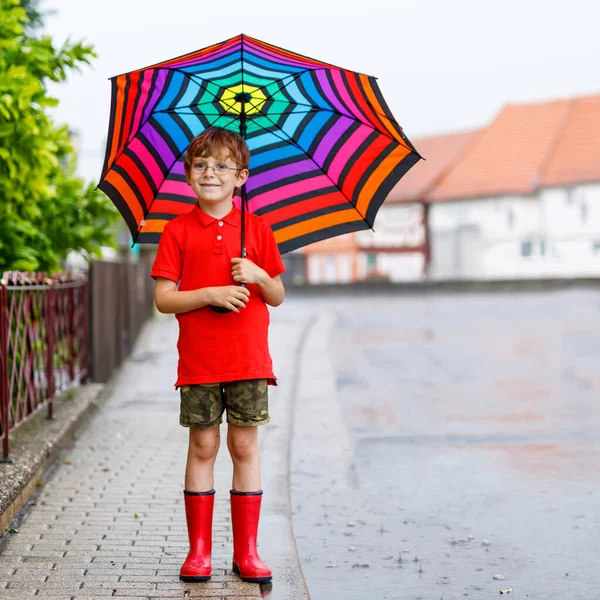 Menino feliz vestindo botas de chuva vermelha e andando com guarda-chuva colorido na rua da cidade. Criança com óculos no dia de verão. criança feliz durante chuva forte do chuveiro do verão — Fotografia de Stock