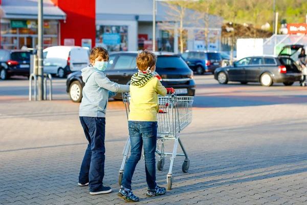 Twee jongens met een medisch masker als bescherming tegen een pandemische coronavirusziekte. Kinderen die beschermende uitrusting gebruiken tegen covid 19 en gaan winkelen in de supermarkt met karretje trolley. — Stockfoto